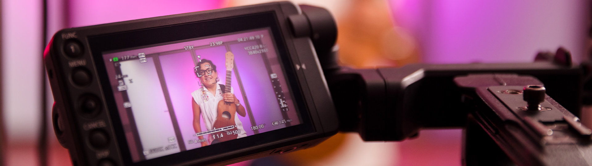 Photo of a camera viewfinder and video set up. In the viewfinder a woman can be seen holding a guitar and speaking.