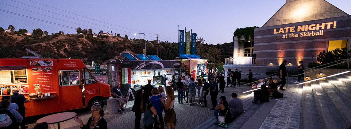 Photo of the front steps of the Skirball. People are standing around talking and ordering food from parked trucks. The words "Late Night!" are projected in lights on the side of the building.