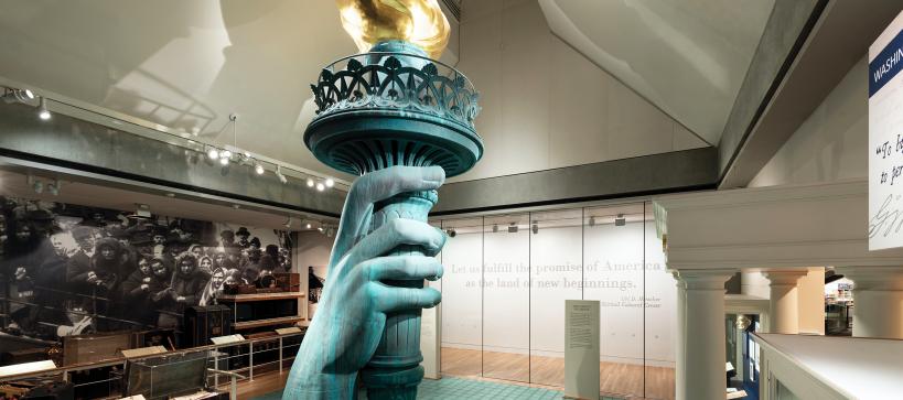 Photo inside the exhibition Vision and values showing a replica of the statues of liberty hand holding the torch among a large photo mural of immigrants on Ellis Island