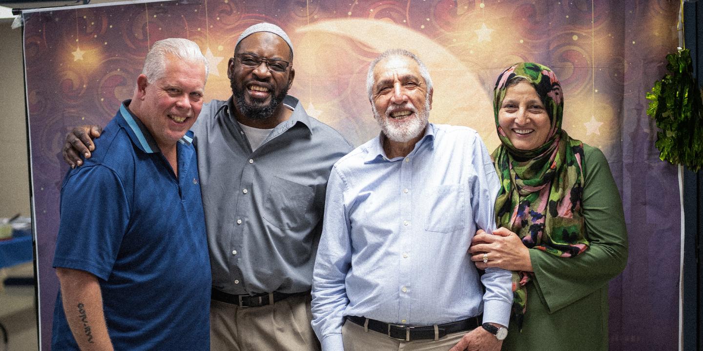 Photo of four people posing in front of a colorful backdrop. Three men and a women wearing a headscarf are all holding each other and smiling at the camera