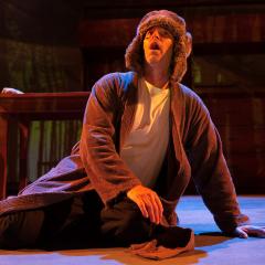 Photograph of a man kneeling on a stage looking up. He is dressed in a long, brown robe and wearing a hat that looks like floppy dog ears.