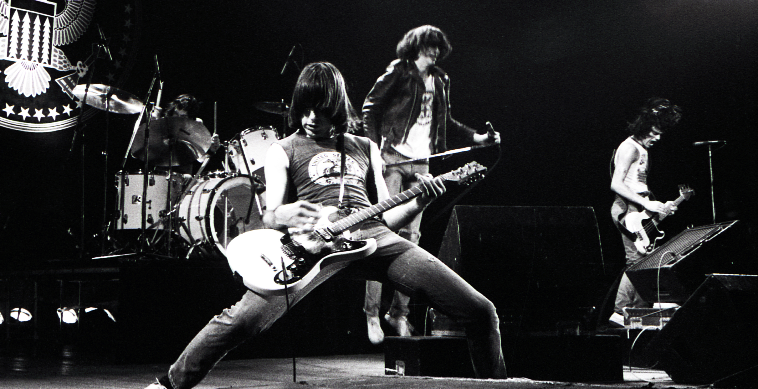 A black and white photo of a guitar player up front from his band on a stage playing.