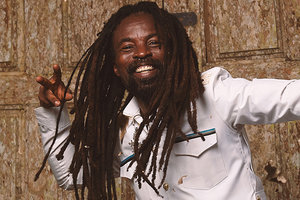 Photo of Rocky Dawuni wearing a white suit and dancing in front of a door. His arms are outstretched and brown leaves are falling around him.