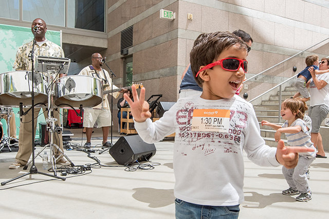 Photo of a Family Amphitheater Performance at the Skirball. A band plays their instruments on the left of the frame and children can be seen dancing around them. In the center is a young child in sunglasses jumping up and down smiling.