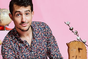 Headshot of Guy Mintus sitting in front of a pink background and surrounded by various, whimsical toys and items