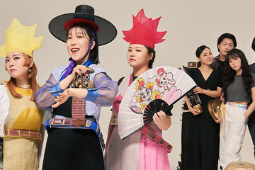 ADG7 band members standing against a blank wall. Three members have colorful clothing, hats, and fans and the other members are standing behind them in gray holding instruments