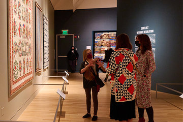 Image from inside a gallery space in Fabric of a Nation: American Quilt Stories. Large quilts are hanging on all walls and three women stand in the center of the frame discussing a quilt in front of them.