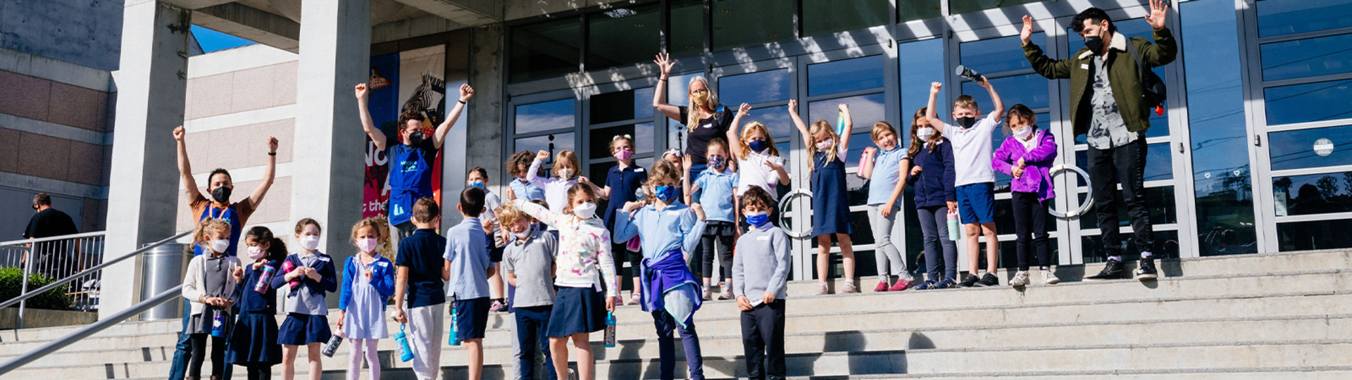 Photo of a school group of children standing on the steps of the Skirball Cultural Center. They are all wearing face masks and waving at the camera.