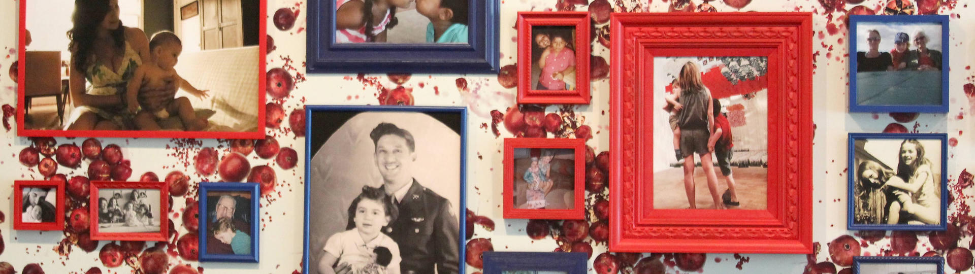 Various photographs of people framed in red and blue frames on a wall covered in wallpaper with a pattern of pomegranites