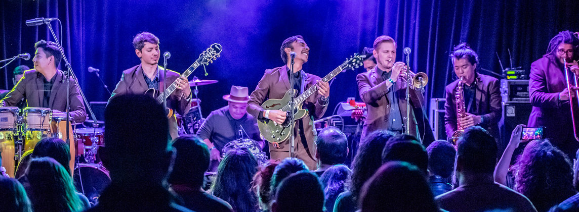 A group of seven musicians playing on purple and blue-lit stage in front of an audience of people standing