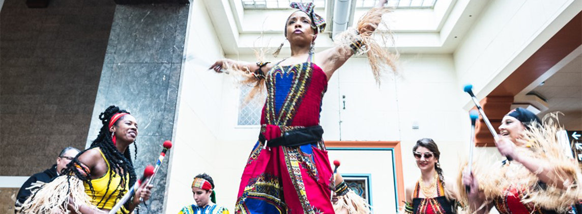 Photo of the members of Bloco Obini posed in costume inside a brightly lit room. They are dancing and playing instruments.