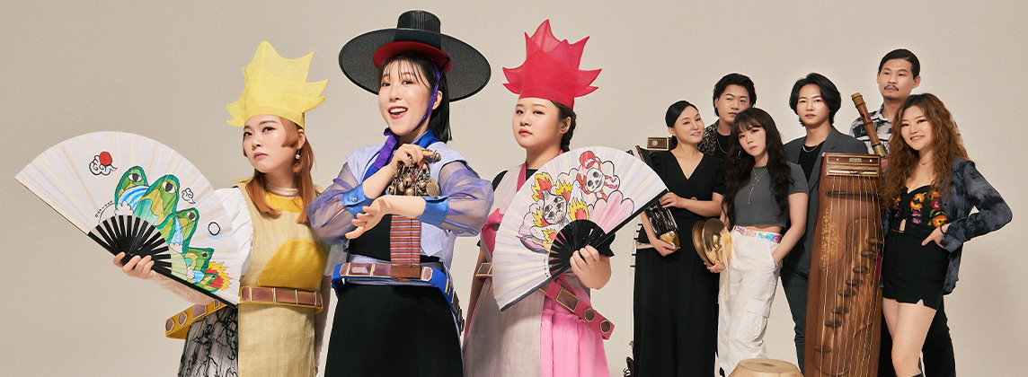 ADG7 band members standing against a blank wall. Three members have colorful clothing, hats, and fans and the other members are standing behind them in gray holding instruments