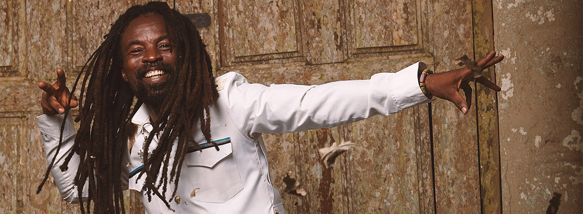 Photo of Rocky Dawuni wearing a white suit and dancing in front of a door. His arms are outstretched and brown leaves are falling around him.