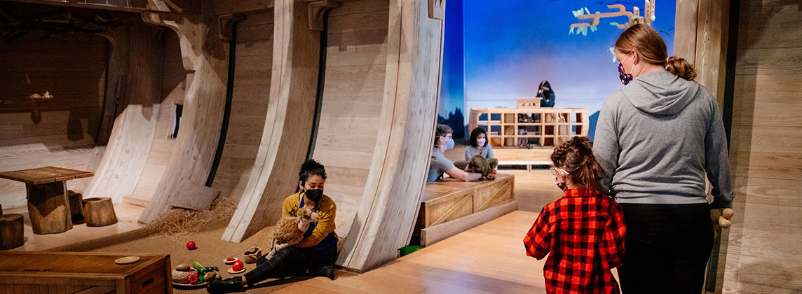 An adult and child stand inside Noah's Ark and watch a masked puppeteer interact with the space. More masked individuals can be seen through an opening in the Ark.