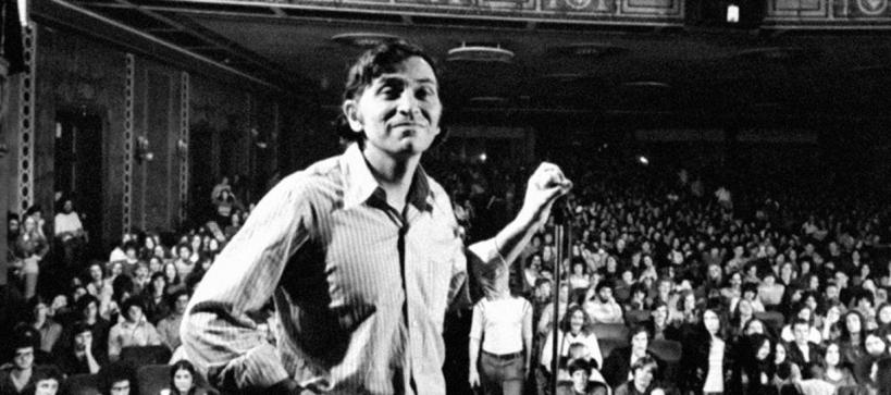 a man on a stage holding a microphone stand with a large crowd of people in the seats below the stage