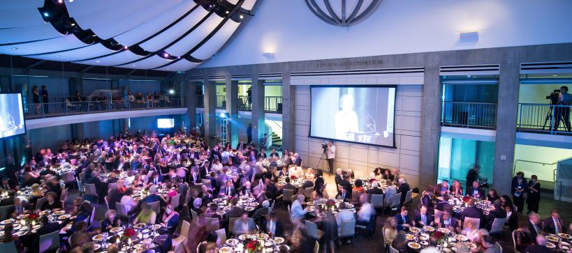 An wide image of an event with people seated at tables eating as they look up to a large screen.