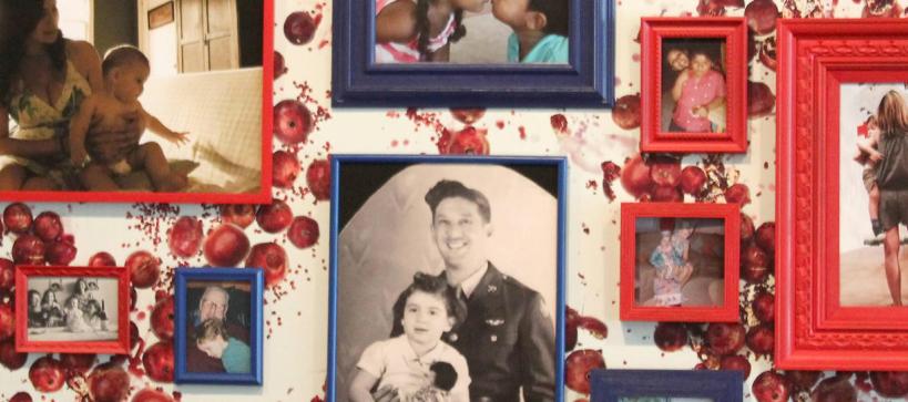 Various photographs of people framed in red and blue frames on a wall covered in wallpaper with a pattern of pomegranites