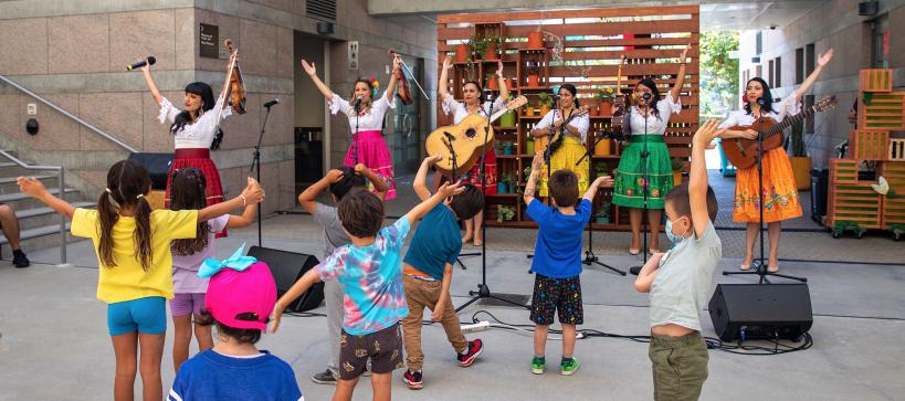 Kids dancing and watching a band of six women playing guitar and wearing brightly colored skirts