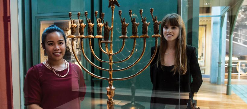 Two visitors looking at a gold Hanukkah lamp with the statue of liberty in the gallery