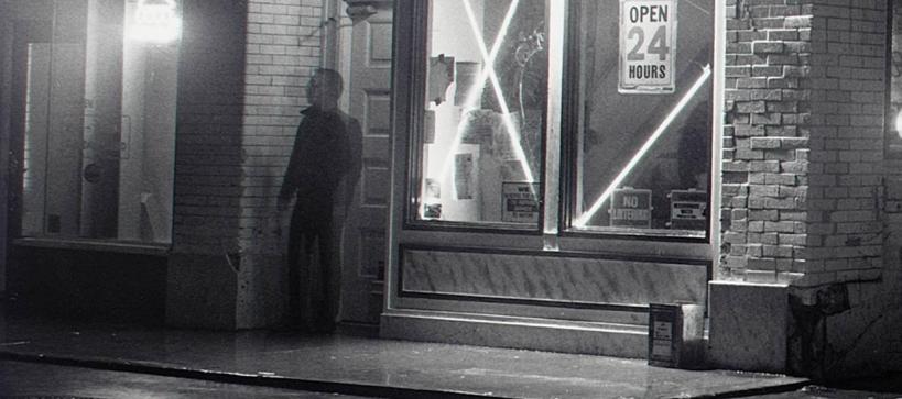 Black and white photo of shadowy man standing outside the doorway of a store at night
