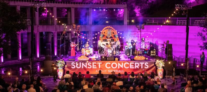 Image of outdoor stage with band playing and audience seated and listening. A large banner on the stage reads Skirball Stages