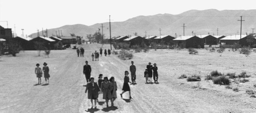 people walking on a dirt road with barracks visible behind them in the distance and mountains even further in the distance