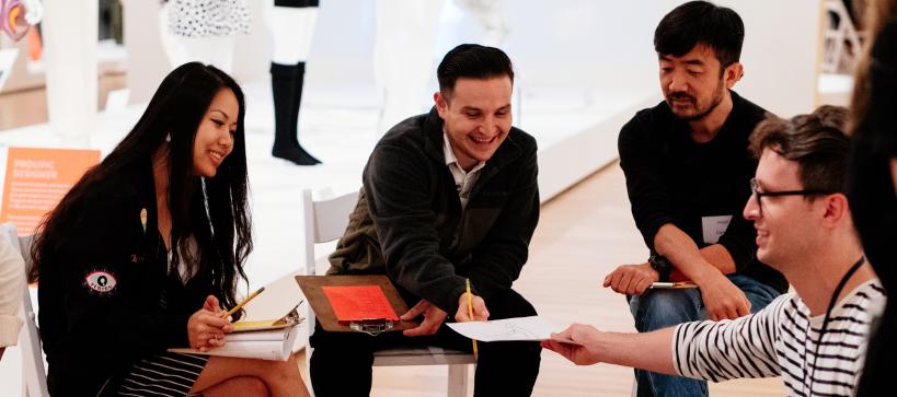 Four adults with clipboards and pencil in a circle looking at a drawing in a gallery.