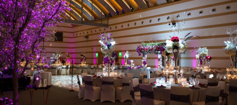 A large hall with table decorated with large vases and purple flowers set up for an event. Dramatic purple lighting fills the banquet hall.