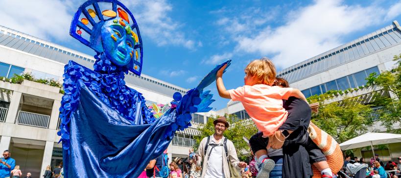 Outdoor photo at the Skirball campus. A large, blue puppet is on the left, reaching out to a young child in their adult's arms and is reaching back. People can be seen behind the trio dancing.