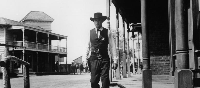 Still from the movie showing Gary Cooper walking towards the camera wearing dark clothing and a cowboy hat. He is surrounded by western buildings and a bright sky.