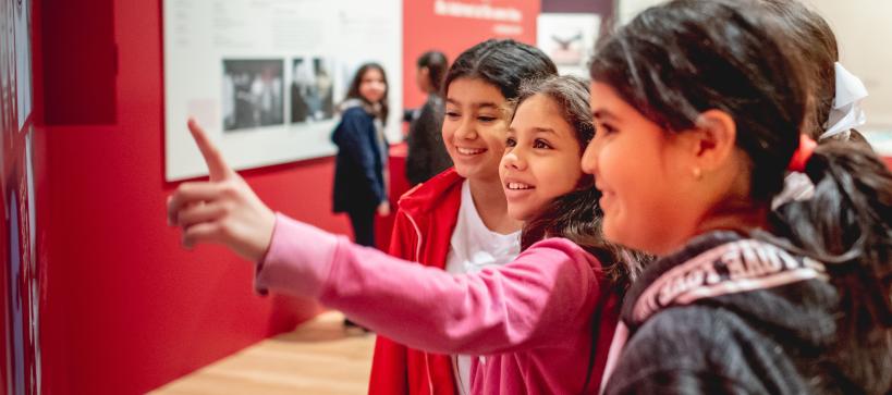 Three young girls are standing together in a red gallery looking and pointing at something out of frame.