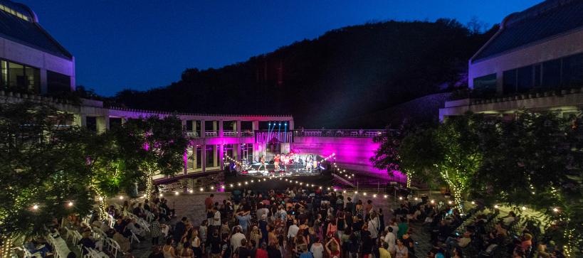 Photo of an outdoor concert at the Skirball. A large group of people are in the courtyard dancing to a band on stage. Lights are seen in the trees, strung across the courtyard, and from the stage.