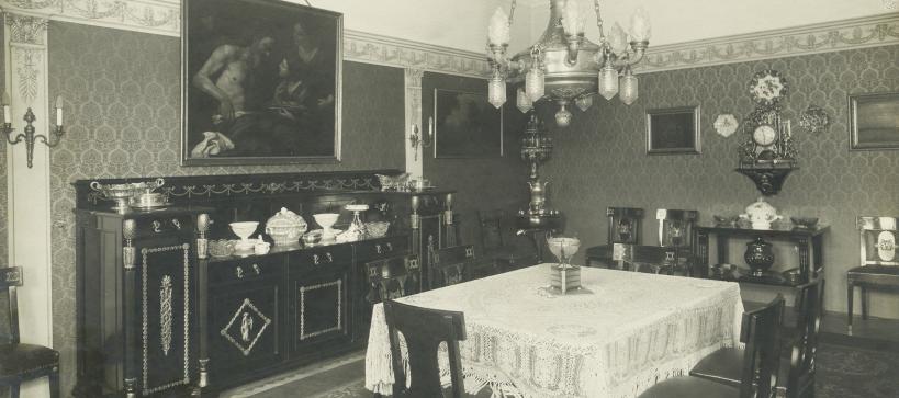 Black and white photo of a dining room with a large table and chairs in the center. On the far wall, a large painting is seen hanging.