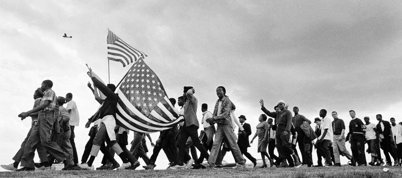 Black and white photo of a large group of people walking on a dirt road from right to left. Two people to the left side of the image are holding American flags.
