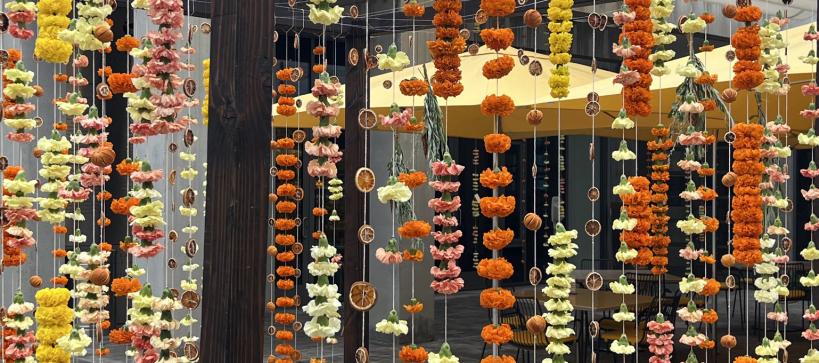 Strings of yellow and orange carnations and mums, and orange slices hung vertically on a wooden structure creating a cozy shelter for a table and chairs.