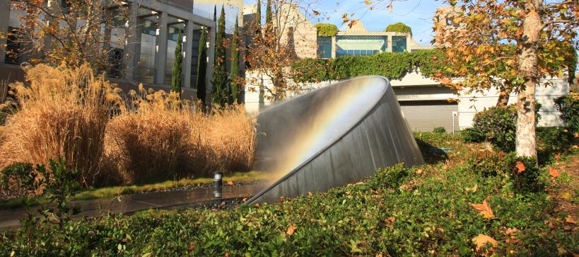 A metal sculpture that shaped in an arc emits mist and creates a rainbow. The sculpture sits in an green arroyo outside of a 3 story building.