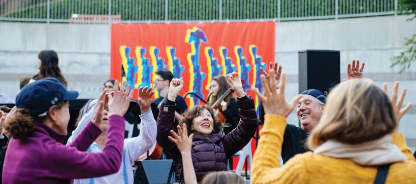 A group of people dancing outside in front of a large banner of a hanukkiah