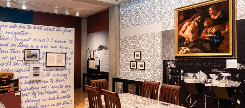 In a gallery with a dining room with a large table and chairs in the center. On the far wall, a large painting is seen hanging.