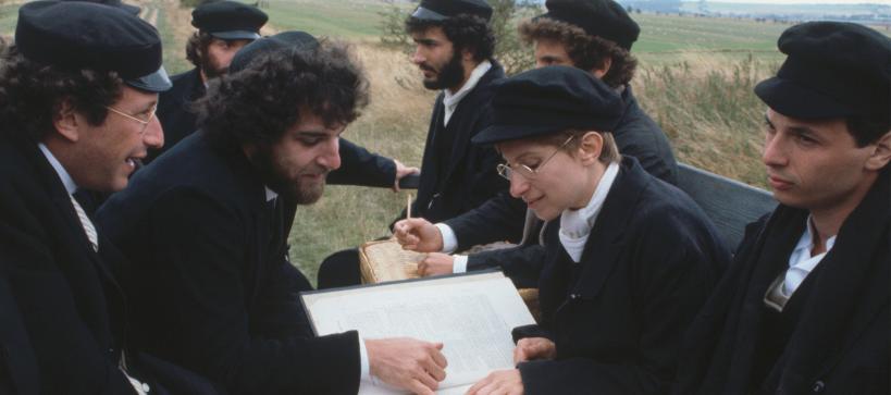 Still from the movie showing Mandy Patinkin and Barbra Streisand sitting with other men in the back of a truck reading religious text