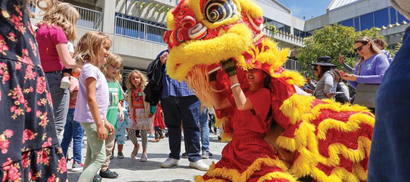 In a large outdoor crowd, two young kids stand before a performer operating a large red and yellow dragon puppet