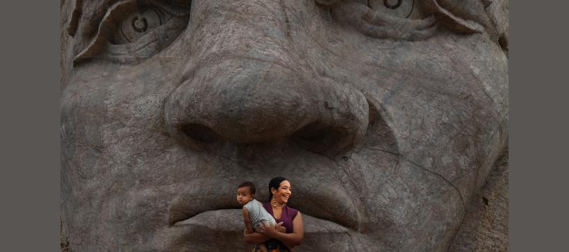 A woman holds a young child in front of a large stone sculpture of a human face.