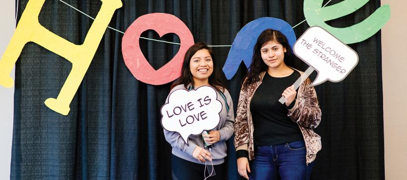 two girls holding speech bubble signs that read, "love is love", and "welcome the stranger".