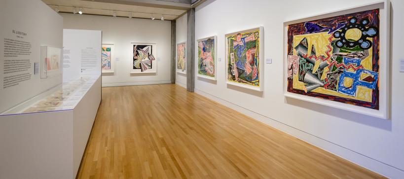 A gallery filled with large, colorful artwork in frames faces a long counter-like display of prints on paper.