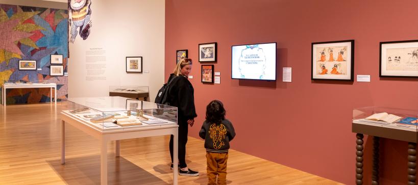 An adult and child look at a screen on a red gallery wall. Around them are framed artworks, tables displaying open books, and a large illustration of a monster seeming to hang from the top of a wall.