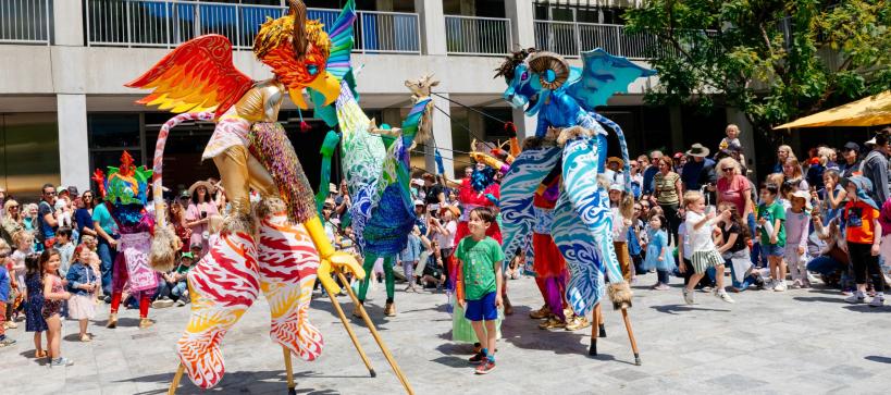 Performers on stilts in elaborate costumes move and dance in the middle of a large group of families in a Courtyard.