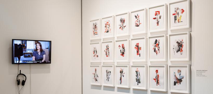 Photo of a gallery corner with a flat screen displaying a colorful video next to 18 framed collages hung in a grid.