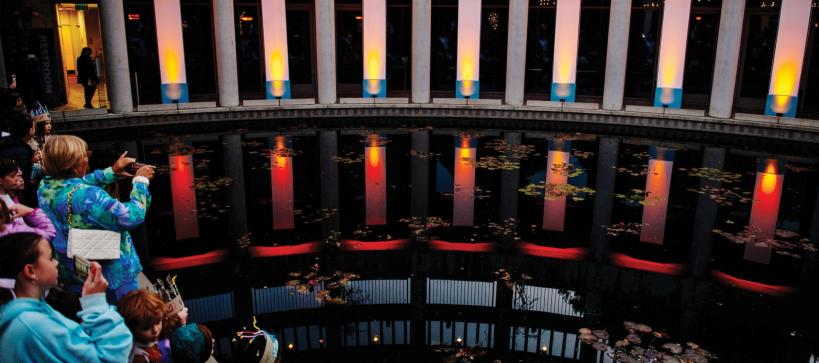 Photo of the lily pond at the Skirball lit up with large column spotlights mimicking a Hanukkah. Families stand along the pond looking at the lights.