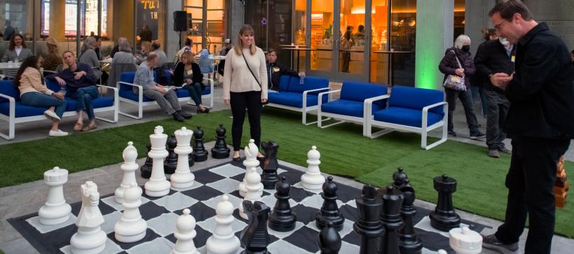 Two people are playing a giant chess game on a courtyard full of people.