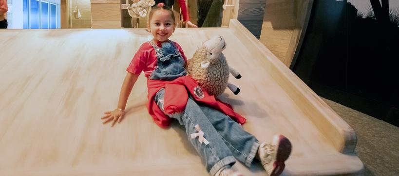 I girl slides down a wooden slide holding a stuffed sheep, smiling.