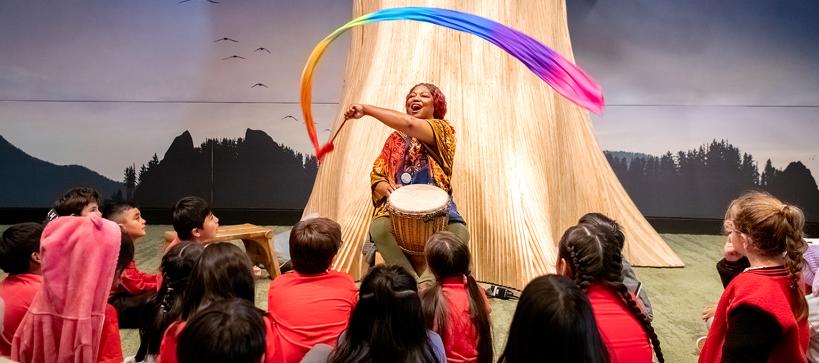 An educator waves a rainbow ribbon playing a drum for kids in a gallery.